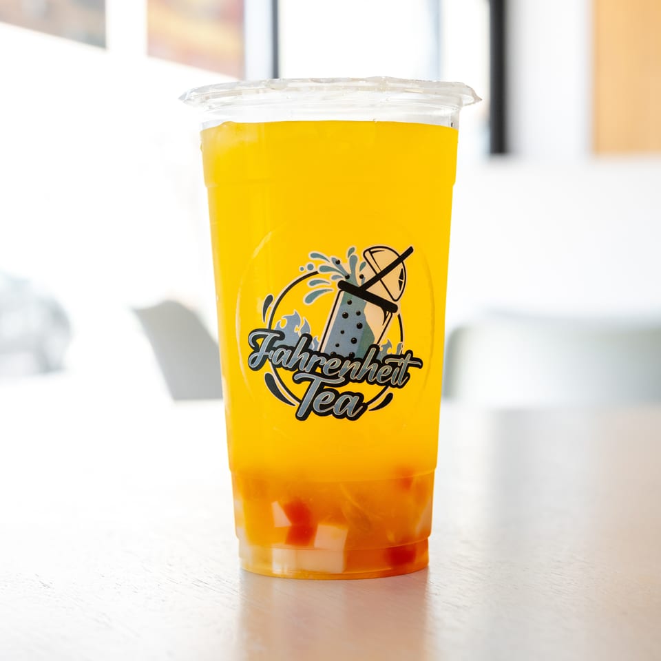 A Fahrenheit Tea drink, a vibrant yellow beverage with fruit pieces, sits on a light wooden table.