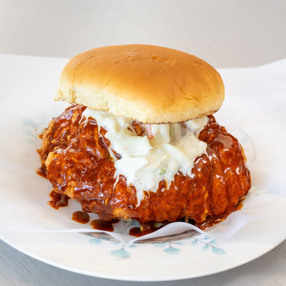 Spicy fried chicken sandwich with coleslaw and sauce on a white plate with floral accents.