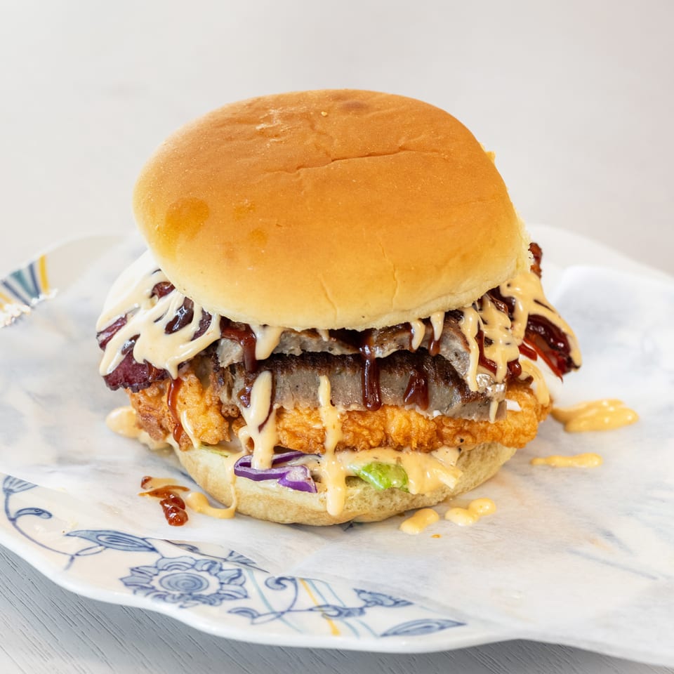 Loaded burger with a crispy fried chicken fillet, beef patty, bacon, lettuce, onions, and drizzled sauces on a patterned plate.