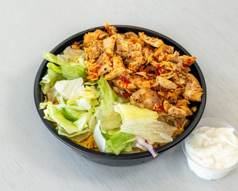 Chicken and rice bowl with lettuce and a side of white sauce on a white table.