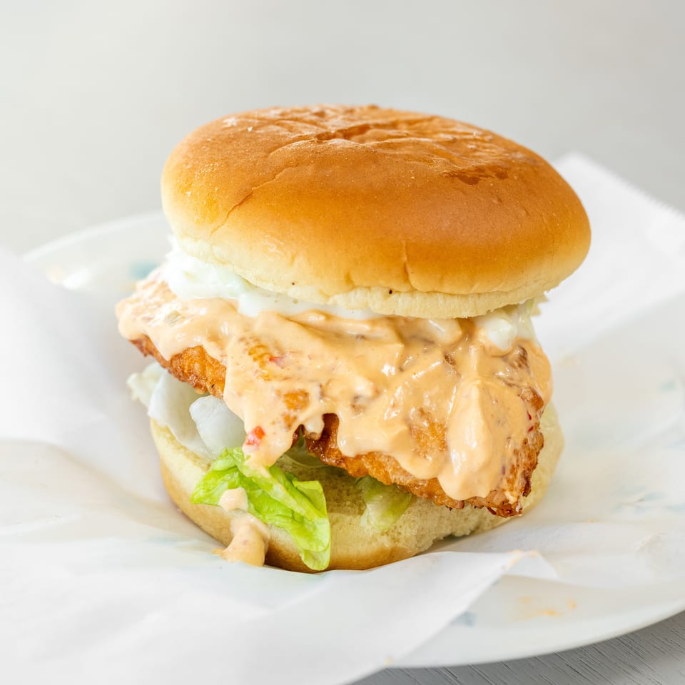 A fried chicken sandwich with lettuce, white sauce, and spicy orange sauce on a bun.