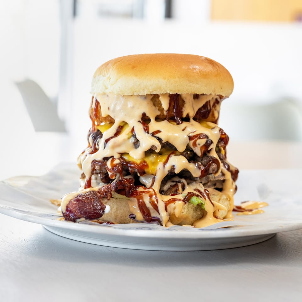 A towering triple-patty burger with melted cheese, bacon, and generous drizzles of sauce on a white plate.