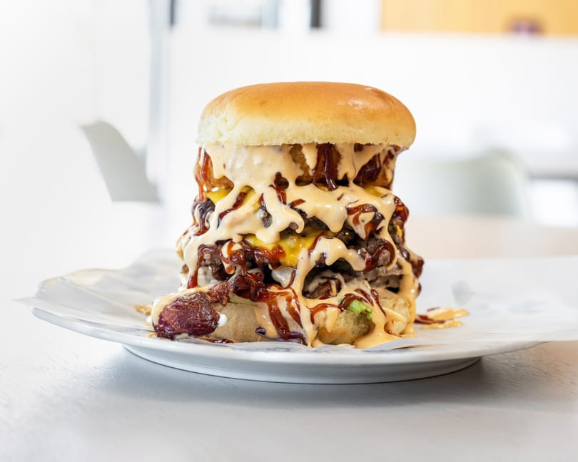 A towering triple-patty burger with melted cheese, bacon, and generous drizzles of sauce on a white plate.