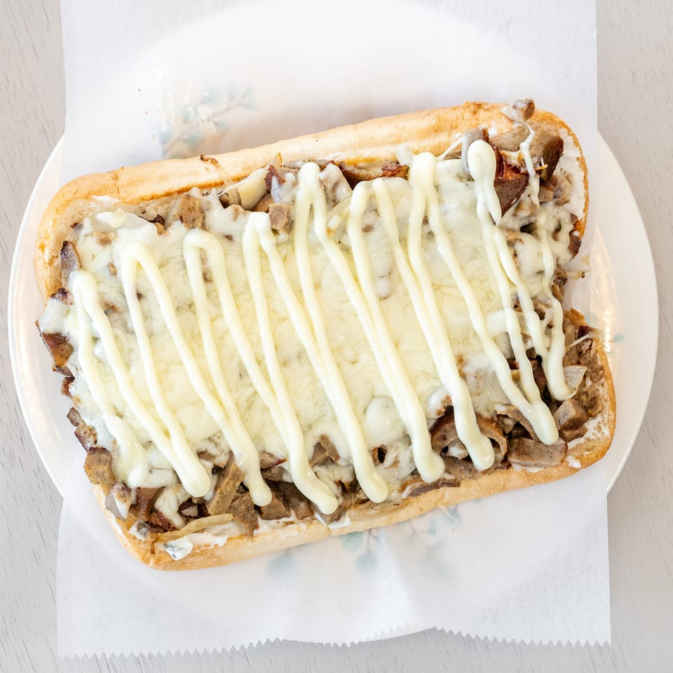 Toasted steak and cheese sandwich topped with melted cheese and creamy sauce on a white plate.